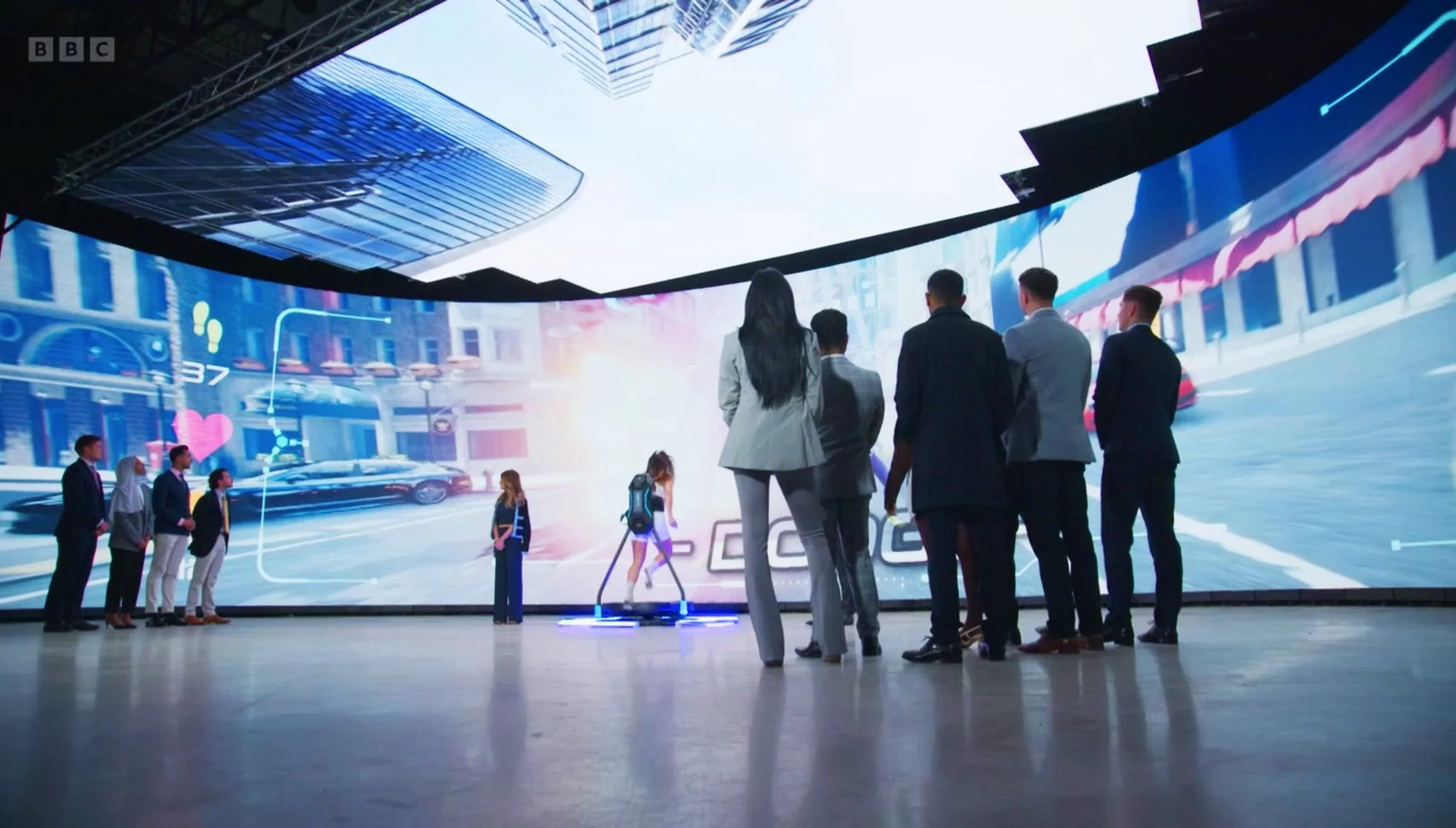 Visitors watching an interactive 3D city environment on a large curved screen, with one person using a motion platform controller